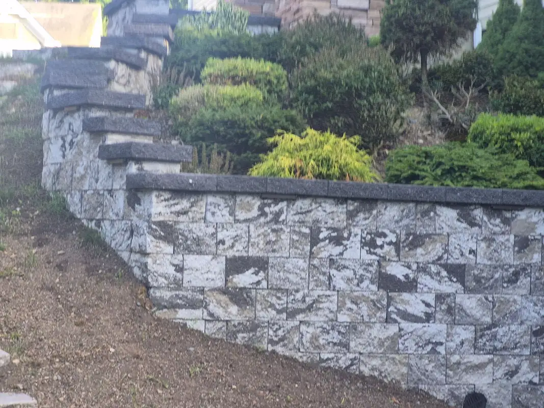 Stone retaining wall with pillars and landscaping near a driveway.